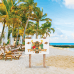 A photo of a tropical beach along the ocean. There's sand, palm trees, blue sky and wooden reclining beach chairs. There is a white sign on wood pols. The sing reads "Tiki Bar" with a picture of a tiki on it. The sign was printed on GF 201HTAPAE.