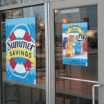 A photo of a glass door and large window at the front of a store. There are two blue static cling signs attached to the windows. One says, " Summer Savings" in black text on top of a live preserver, the other has a photo of some beverages for $2.99. The static clings are printed on GF 208.