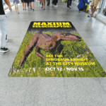 A photo of a hallway with a vinyl graphic adhered to the ground. It has a CGI image of a velociraptor with the text, "Maximum Velociraptor!" written above it in large yellow letters. Below, there is smaller yellow text that says, "See the dinosaur exhibit at the City Museim," as well as white text the same size that says, "Oct 12 - Nov 15." This graphic is printed on GF 211.