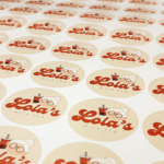 A photo of circular yellow stickers on a sheet that say, "Lola's" in a large red cursive font and, "MILK TEA" in smaller black all caps underneath. There is an illustration of an elderly woman with white hair and brown circular glasses with her hair up in a bun holding a cup with a straw. These stickers were printed on GF 214.