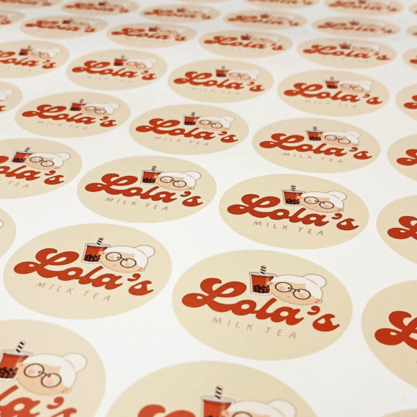 A photo of circular yellow stickers on a sheet that say, "Lola's" in a large red cursive font and, "MILK TEA" in smaller black all caps underneath. There is an illustration of an elderly woman with white hair and brown circular glasses with her hair up in a bun holding a cup with a straw. These stickers were printed on GF 214.