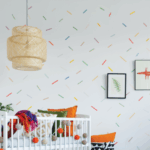 Photo of a baby's nursery. There is a white crib with orange, green and white pillows and a purple and orange blanked. There is a white, orange, and green garland of plush spheres on the outside of the crib. Hanging from the ceiling is a light inside a tan, whicker lampshade. There are framed images of a fern and and orange dog on the wall. The Wallpaper is white with different colored lines, mimicking sprinkles on a cake. The Wallpaper is printed on GF 226.