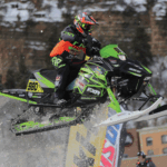 A photo of a green and black snowmobile ridden by a rider going off a jump in a competition. There are various decals on the snowmobile printed on GF 235.