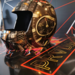 A photo of what appears to be an old helmet that covers the whole head, leaving a space for a person's face to show. The helmet is black and covered with a gold design that looks like a closeup of a watch. The surface the helmet is sitting on as well as the wall behind it are black with abstract designs in red, pale blue, yellow, and gold. Used to advertise GF 242.