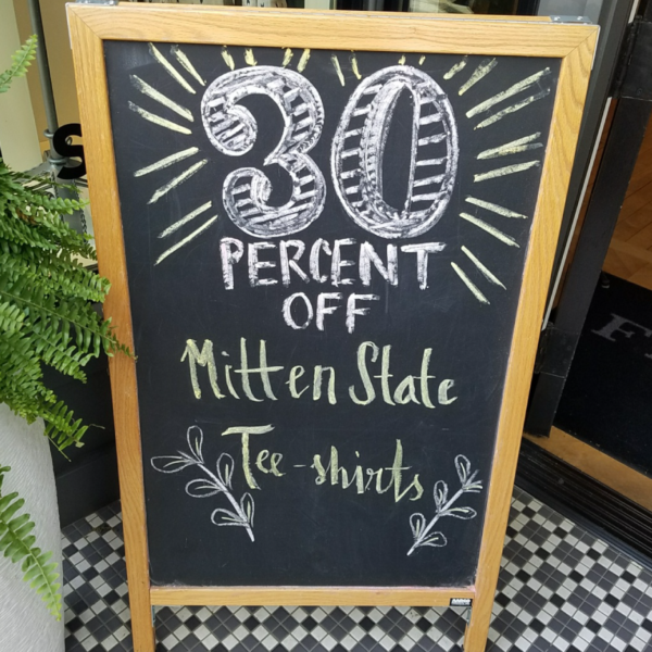 A photo of a black chalkboard sign on a wooden, A-frame stand. Someone has written "30 percent off Mitten State Tee-shirts," using white and green chalk. The chalkboard is GF 290.
