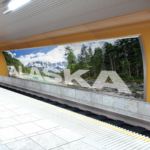 A photo of a subway station with a large advertisement on the wall across the tracks. The ad consisting of a photo of a forest, river, and mountains, with," ALASKA," writing in large, white all-caps with slightly lower opacity. The image was printed on GF 275.
