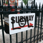 A photo of a white sign that reads, "SUBWAY," in black, sans-serif all caps with a red arrow pointing to the right. The sign is attached to a black, iron fence with spikes on the top. Someone has spay painted graffiti that reads, "LOL," in all caps with a smiley face in the O. Used for GF 108 sales listing.