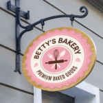 A photo of a circular sign hanging on a decorative black iron hood that. The sign is pink, white, and tan, and in black text reads, "Betty's Bakery - Premium Baked Goods," in black, all caps text. The text is set in a circle shape on white. In the middle is a pink circle with illustrations of a rolling pin, whisk, fork, and spoon. The sign is printed on GF 201OPAE.
