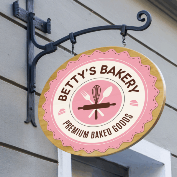 A photo of a circular sign hanging on a decorative black iron hood that. The sign is pink, white, and tan, and in black text reads, "Betty's Bakery - Premium Baked Goods," in black, all caps text. The text is set in a circle shape on white. In the middle is a pink circle with illustrations of a rolling pin, whisk, fork, and spoon. The sign is printed on GF 201OPAE.