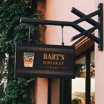 A photo of a black sign hanging off an iron sign holder on the side of a building. In marigold text is reads, "Bart's Whiskey Bar," next to an image of a glass of whiskey. The sign is printed on GF 203OP.