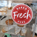 A photo of a clear plastic display with doors containing backed goods on trays. There is a round, red decal on the front that reads, "BAKED Fresh HERE," in a mix of san-serif, all caps text and regular case script font. The decal is printed on GF 216.