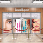 A photo of a storefront with the world, "Boutique," written above the doors in silver, all lower-case font. There are decals on the window containing photos of the faces of two women and the words, "Grand Opening," in all-caps pink text and "This Summer," in all-caps teal text. There is a red barrier rope on red stands in front of the door. The window decals were printed on GF 218.