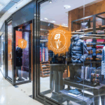 A photo of a clothing store in a shopping mall. There are circular orange decals on the windows with an illustration of a main in a suit and fancy hat in white inside a circle. On the outside there is white text that reads, "Donovan's Men's Clothing. The decals were printed on GF 219.