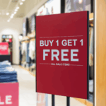 Photo of a red sign in a black holder that reads "Buy 1 get 1 free," in white text. It is standing in the entrance of a shop. Used for product GF 162
