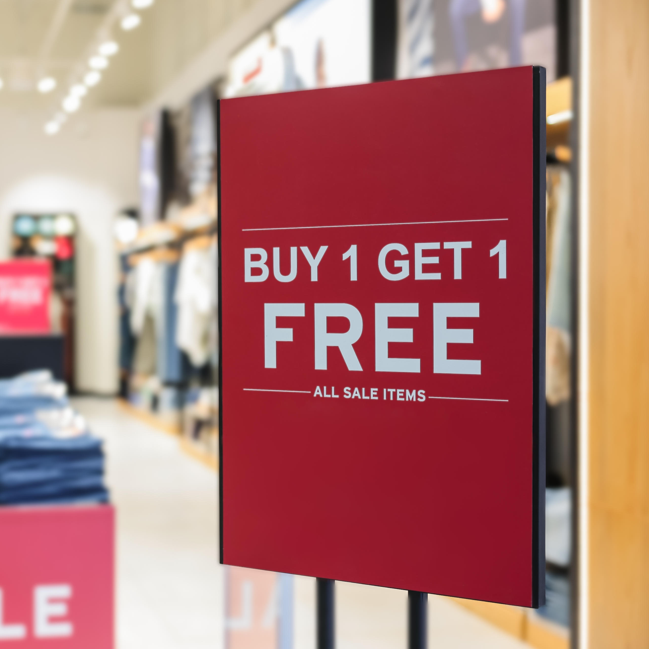 Photo of a red sign in a black holder that reads "Buy 1 get 1 free," in white text. It is standing in the entrance of a shop. Used for product GF 162