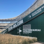 A photo of a sports stadium with a focus on the side of bleachers. If is a sloped, triangular, green wall covered in the logos and website url of the Texas Farm Bureau Insurance. These graphics are printed on GF 240.