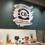 A photo of what appears to be the inside of a restaurant. There is a white wall and counter full of items to buy, and above there is a half-wall dropping down from the ceiling. The wall is green and has a white graphic on it that depicts a burger and text that reads, "Crossroads Plant Based Burgers and Sausages." The graphic was printed on GF 246.