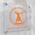 A photo of a grey wall with four metal clips attached to the wall. The clips are holding a piece of clear glass with an orange graphic on it. The graphic is a circle outlined with various patterns and an ancient greek man aiming a bow and arrow. Below is orange text that reads, "Ancient Greece Exhibit," with an arrow pointing to the right. The orange graphic is printed on GF 255.