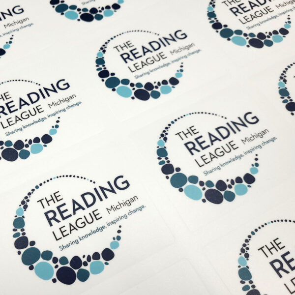 A photo of kisscut vinyl stickers on a sheet. The design on the stickers is circular, made up of multiple circles of various shades of blue with the text, "The Reading League Michigan. Sharing knowledge, inspiring change." These stickers were printed on GF 402.