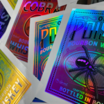 A photo of various rainbow holographic stickers for whisky and bourbon. They contain illustrations of spiders and cobras. These stickers were printed on GF 765.