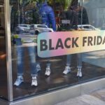 A photo of the front window of a clothing store along a busy street. There are mannequins wearing jeans and winter coats inside. There is a metalic decal with rainbow colors and in large words it reads, "BLACK FRIDAY," in all caps. The decal was printed on GF 364OP.