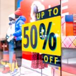 A photo of the front window of a store in a shopping mall. There are mannequins in the window wearing winter clothing, and a black and yellow decal on the window that says, "Up to 50% off." The decal was printed on GF 364OPAE.