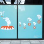 A photo of the windows of a storefront on a sidewalk. There is light-blue vinyl covering the windows with illustrations of white origami elephants riding unicycles with one larger elephant on a circus platform. There is white text in the upper left that reads, "Something new Coming Soon." On the bottom toward the center is smaller white text that reads, "Elephant Park." These images were printed on GF 365OPAE.